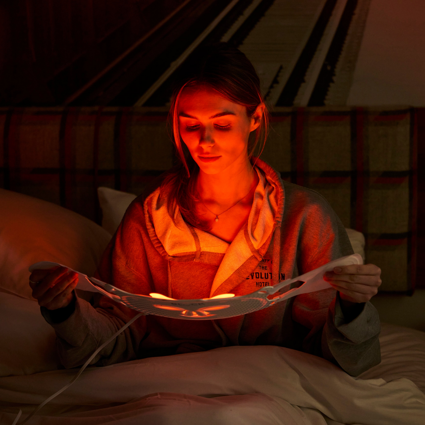 A woman sits in bed reading a red light therapy mask under warm red ambient light in a dim bedroom.