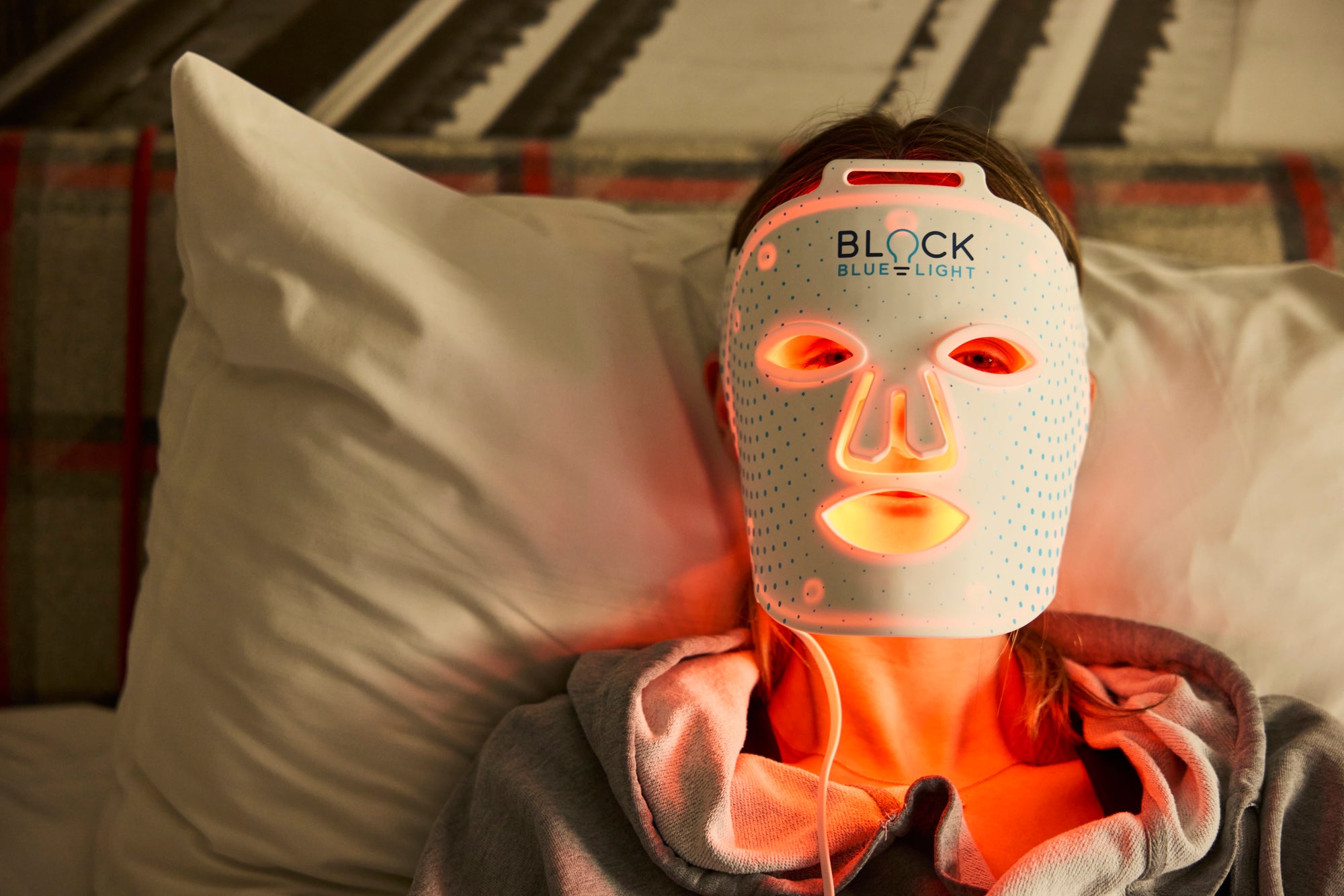 A person lying in bed wearing a white BLOCK Blue Light facial mask that glows orange-red around the eyes, nose, and mouth, with a pillow and blanket visible in the background.