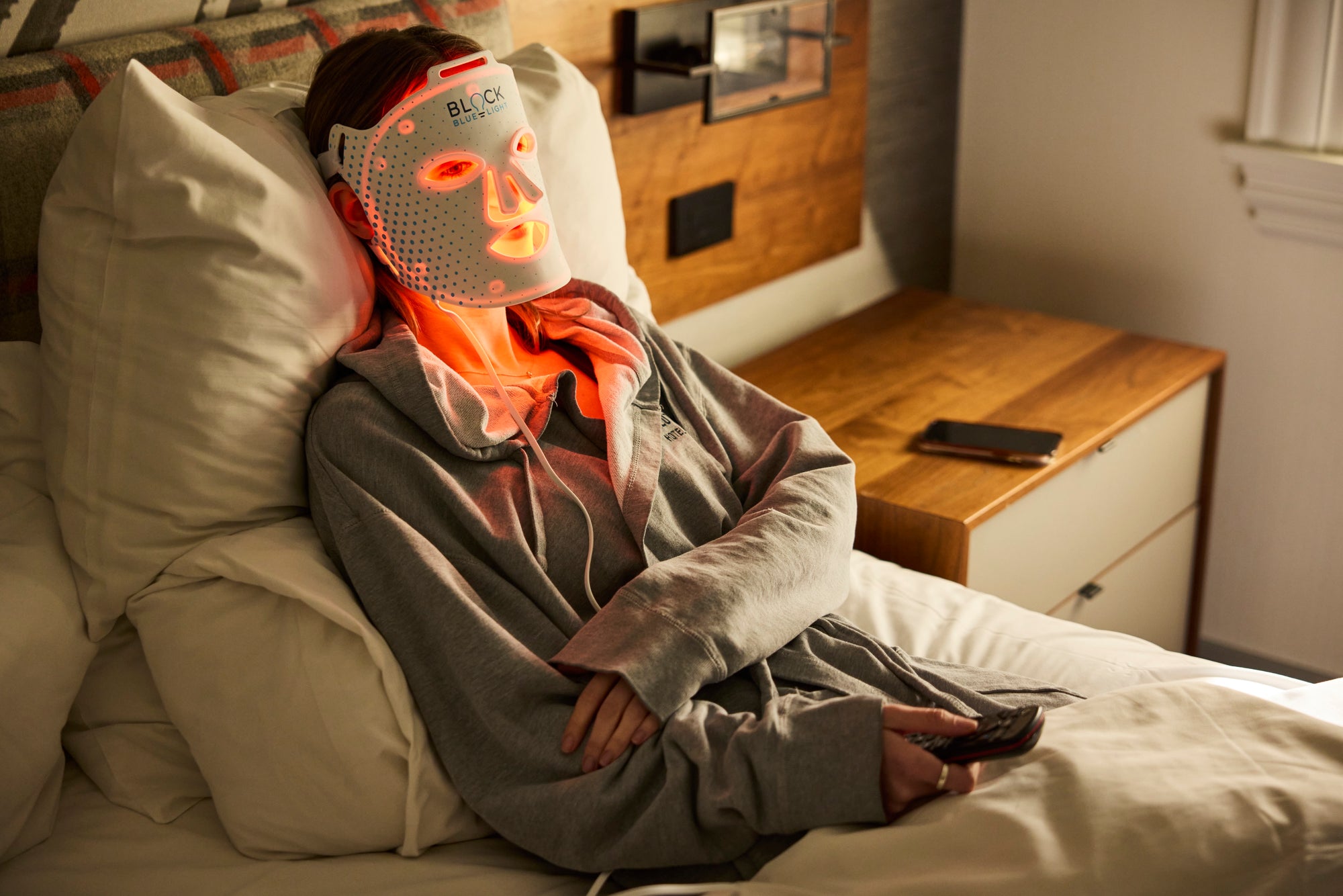 A person reclining in bed wearing a white LED light therapy face mask that glows orange and red, with a gray robe in a bedroom setting.