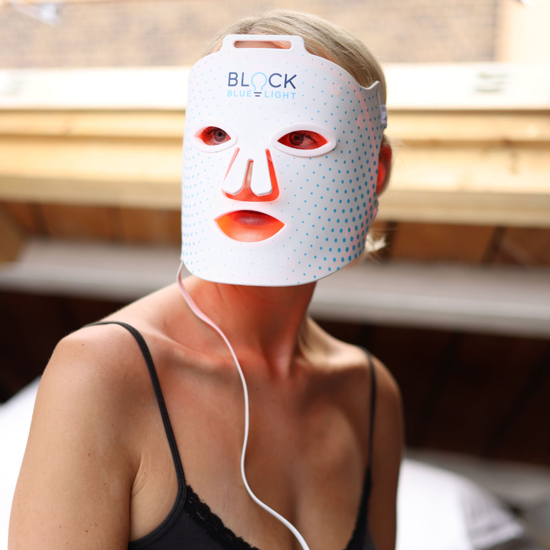 A woman wearing a Block Blue Light red light therapy face mask with blue dots and cutouts for the eyes, nose, and mouth.