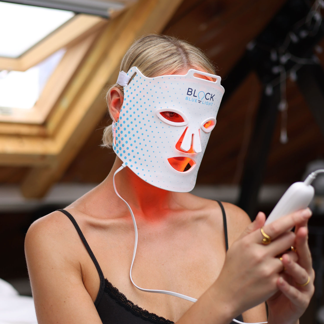 A woman wearing a red light-therapy face mask with blue dotted patterns and an illuminated orange nose opening, holding a controller in a home setting.