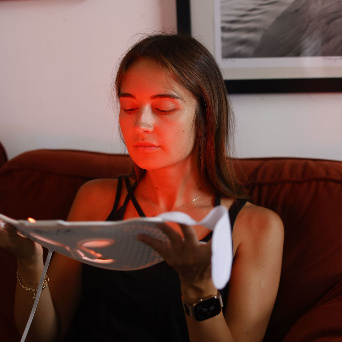 A woman sits on a brown couch holding a Block Blue Light red light therapy mask, with framed wall art in the background.