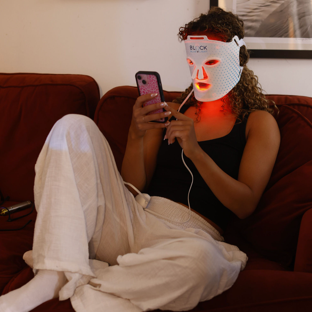 a woman sits on a sofa wearing a Block Blue Light red light therapy face mask.